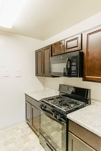 Kitchen with walnut cabinets, white walls, black appliances, tan and white floor. 